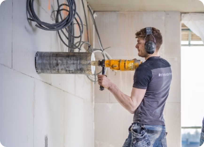 Iemand met een koptelefoon op boort met een grote Thomaboor-boormachine een rond gat in een muur. Boven hem hangen losse kabels en op de achtergrond schijnt zonlicht door een raam naar binnen.