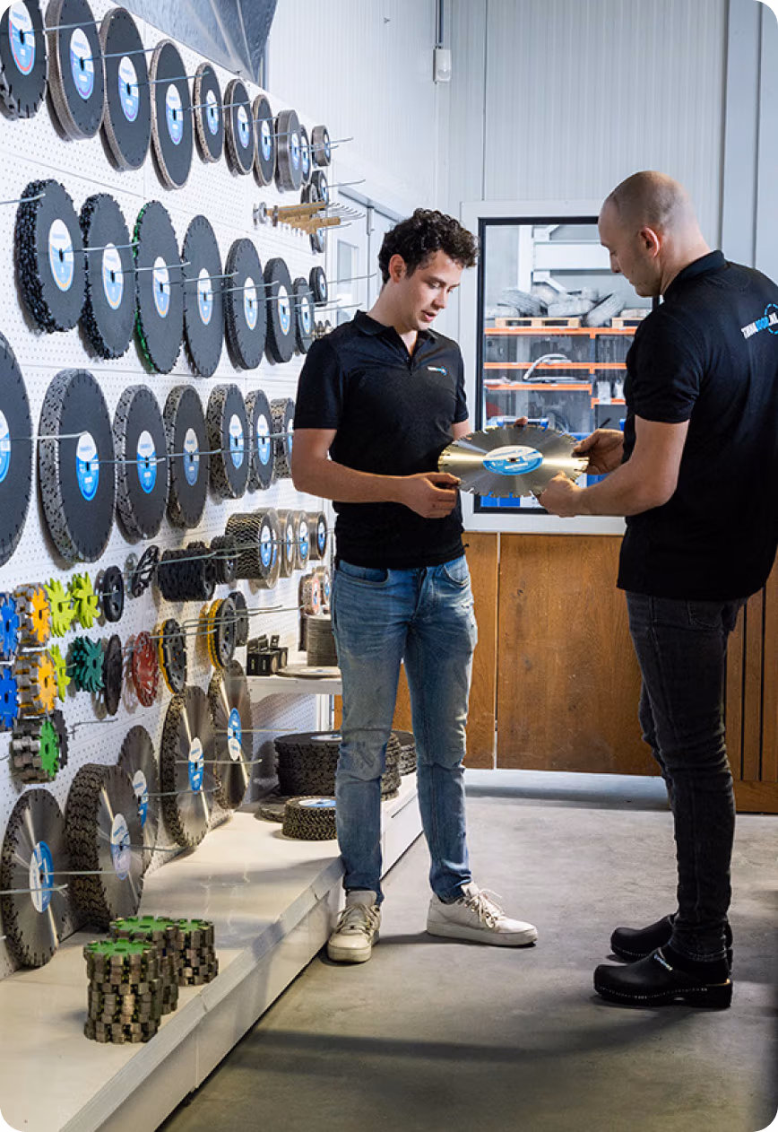 Twee mannen staan in een winkel waar diverse grote ronde slijpschijven, zaagbladen en gereedschappen worden tentoongesteld. De ene man reikt de andere een schijf aan terwijl ze met elkaar praten, omringd door onderdelen van industriële apparatuur, waaronder Diamantboren en Thomaboor-producten.