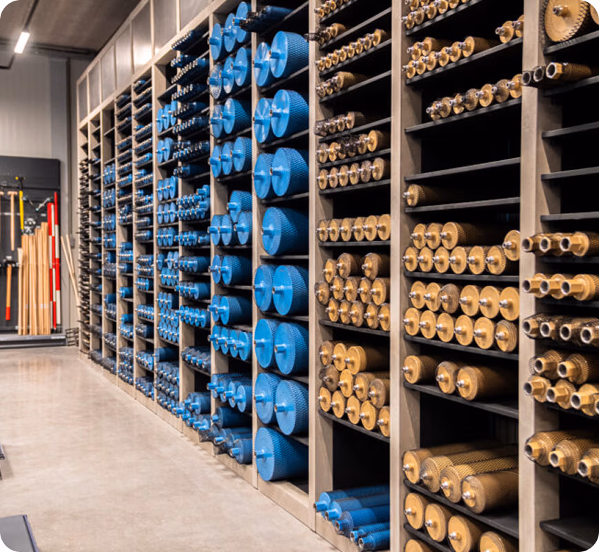Planken vol met netjes gerangschikte metalen rollen, sommige blauw en sommige bronskleurig, die zowel verticaal als horizontaal zijn opgeborgen in een werkplaats of industriële omgeving, naast diamantboren en zaagbladen. Op de achtergrond zijn andere gereedschappen en apparatuur te zien, waaronder producten van Thomaboor.