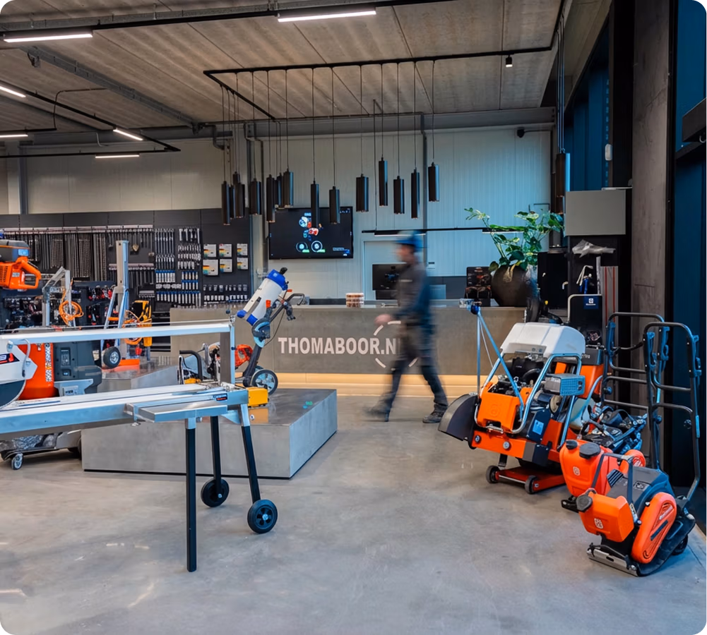 Een moderne showroom met bouwgereedschap en -machines, waaronder zaagbladen en diamantboren, een voorbijlopende persoon en een toonbank met THOMABOOR.NL op de achtergrond. De ruimte heeft betonnen vloeren en een industriële inrichting.