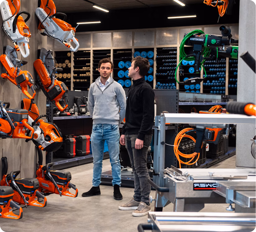Twee mannen staan in een bouwmarkt, omringd door elektrisch gereedschap, zaagbladen en apparatuur die aan de muren en op de schappen zijn uitgestald. Ze lijken te praten over of artikelen te bekijken, zoals Thomaboor-boormachines en diamantboren.