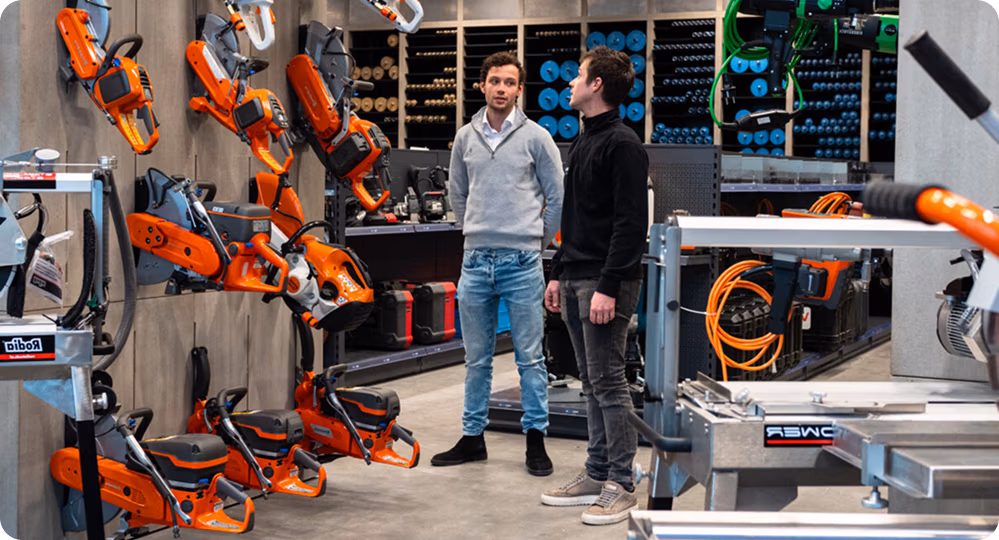 Twee mannen staan in een bouwmarkt en bekijken een display met oranje elektrisch gereedschap dat aan de muur is opgehangen. Naast hen zijn werkbanken, diverse gereedschappen en netsnoeren te zien, evenals schappen vol met hoogwaardige zaagbladen en diamantboren voor professionele projecten.