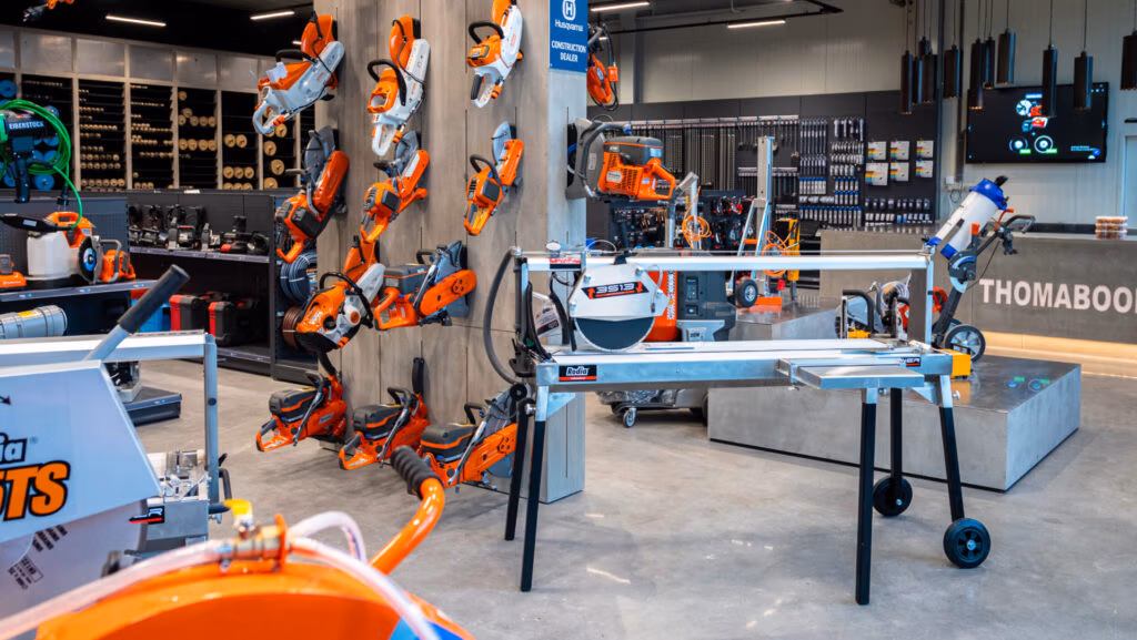 In het interieur van een bouwmarkt liggen oranje en grijze elektrisch gereedschap, waaronder zagen en boormachines, evenals gespecialiseerde apparatuur zoals zaagbladen en diamantboren van Thomaboor, uitgestald op muren en tafels, met op de achtergrond schappen vol accessoires en apparatuur.