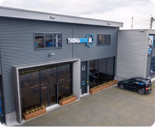 Een modern, grijs industrieel gebouw met grote ramen, een Thomaboor.nl-bord en een kleine zwarte auto die buiten bij de ingang met het nummer 43 geparkeerd staat. Langs de ramen staan rechthoekige plantenbakken, die verwijzen naar de precisie en kwaliteit waarmee de zaagbladen en diamantboren van Thomaboor bekend staan.