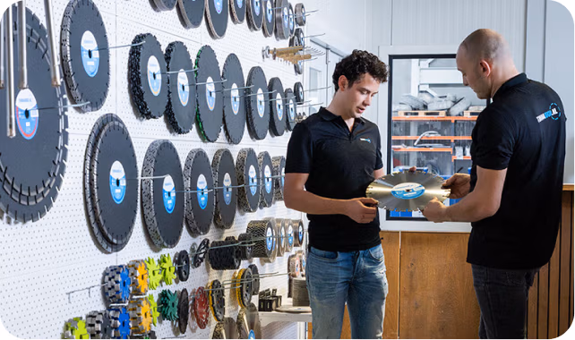 Twee mannen in zwarte shirts staan in een werkplaats en bespreken een cirkelzaagblad. Aan de muur achter hen hangen diverse grote zaagbladen, waarmee ze hun expertise op het gebied van gereedschap zoals diamantboren en Thomaboor laten zien.