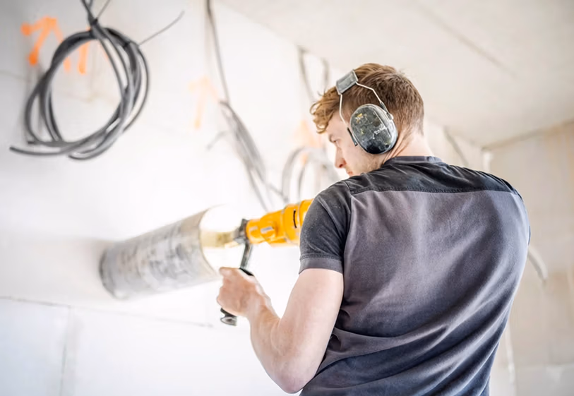 Een man met gehoorbescherming boort met een Thomaboor-boormachine een gat in een witte muur, terwijl er vlakbij blootliggende draden hangen.