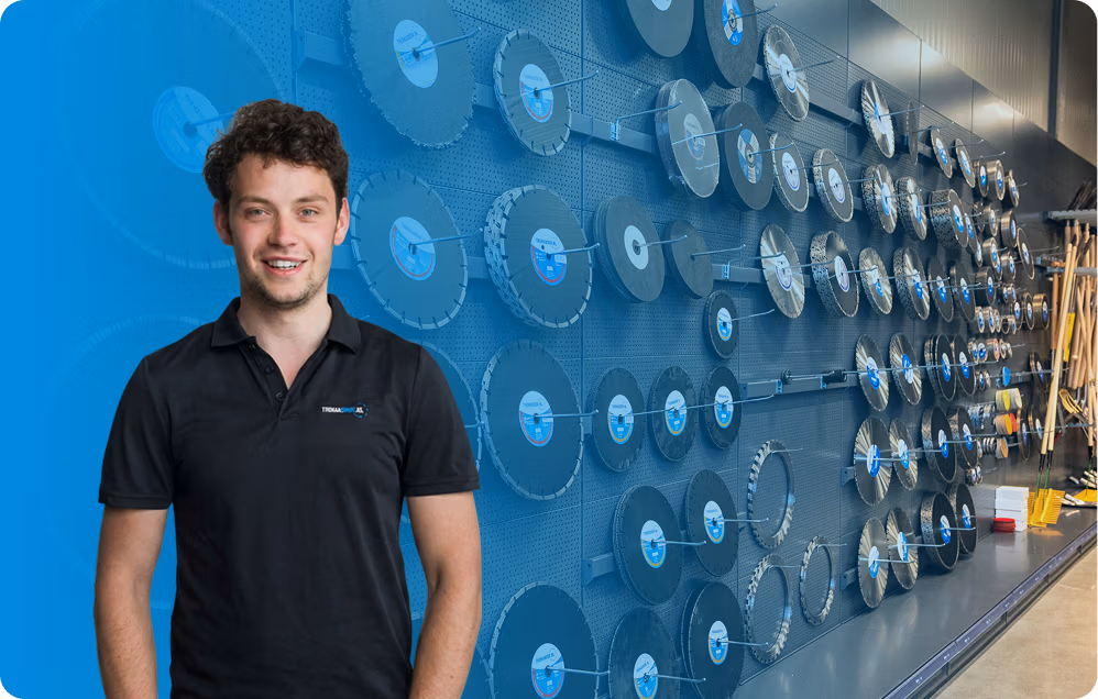 Een glimlachende man in een zwart poloshirt staat voor een muur waarop talrijke grote ronde zaagbladen, waarvan sommige met blauwe etiketten, in nette rijen zijn opgesteld. De indrukwekkende collectie bestaat uit hoogwaardige producten van Diamantboren en Thomaboor.