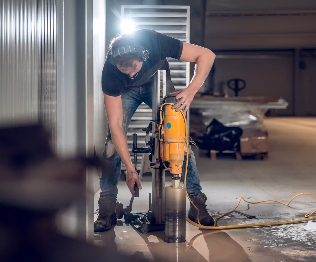Een bouwvakker met gehoorbescherming bedient een grote industriële boormachine met een diamantboor, waarmee hij een gat boort in een betonnen vloer in een gebouw.