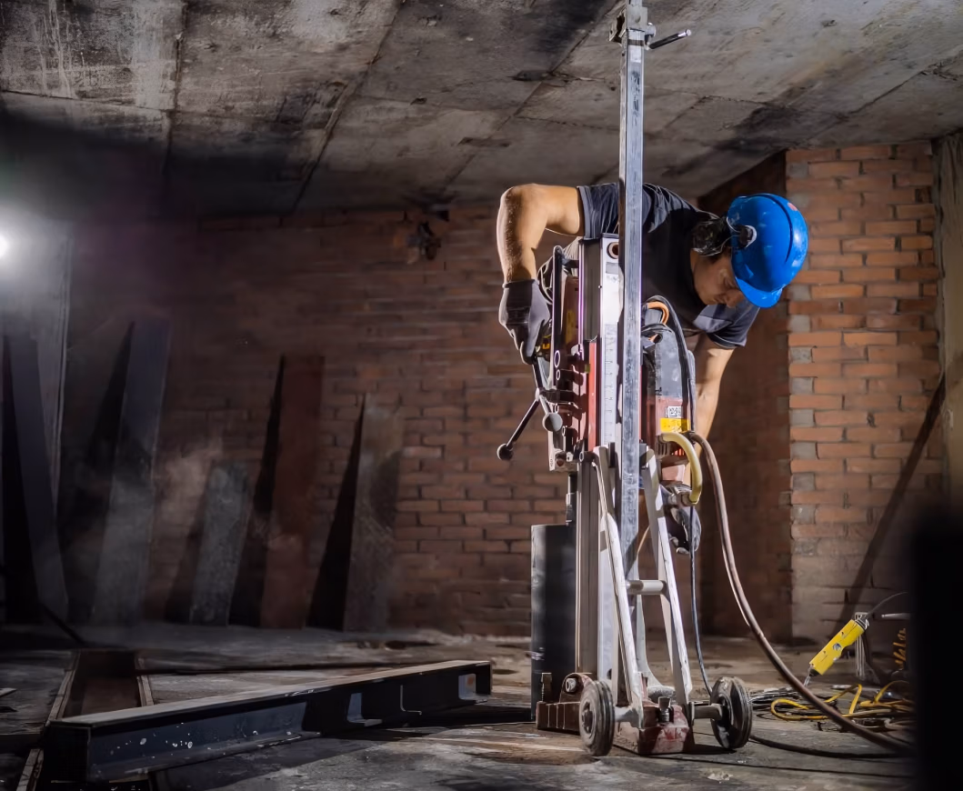 Een bouwvakker met een blauwe bouwhelm bedient zware diamantboorapparatuur op een betonnen vloer in een gedeeltelijk voltooid gebouw met bakstenen muren.