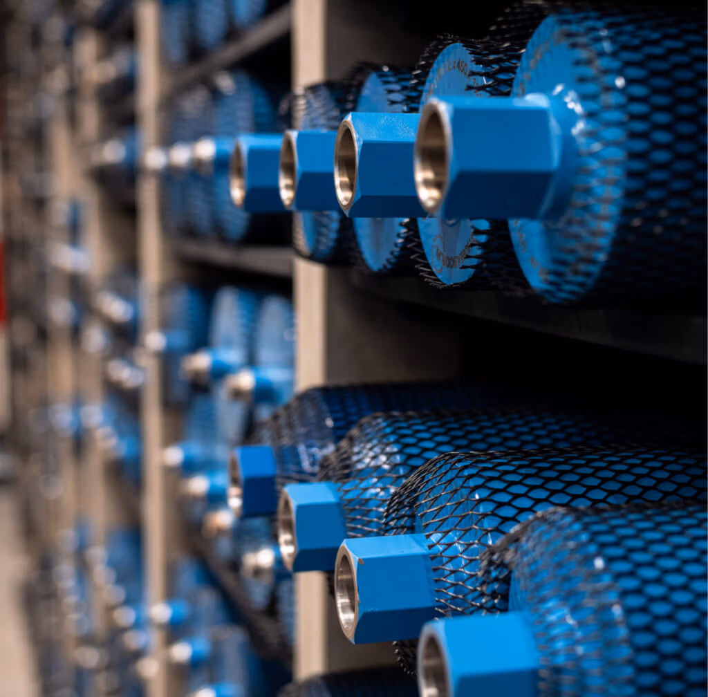 Rijen blauwe, cilindervormige metalen voorwerpen, zoals diamantboren met zeshoekige uiteinden en beschermende gaasafdekkingen, zijn horizontaal op planken gestapeld, waardoor een zich herhalend industrieel patroon ontstaat.