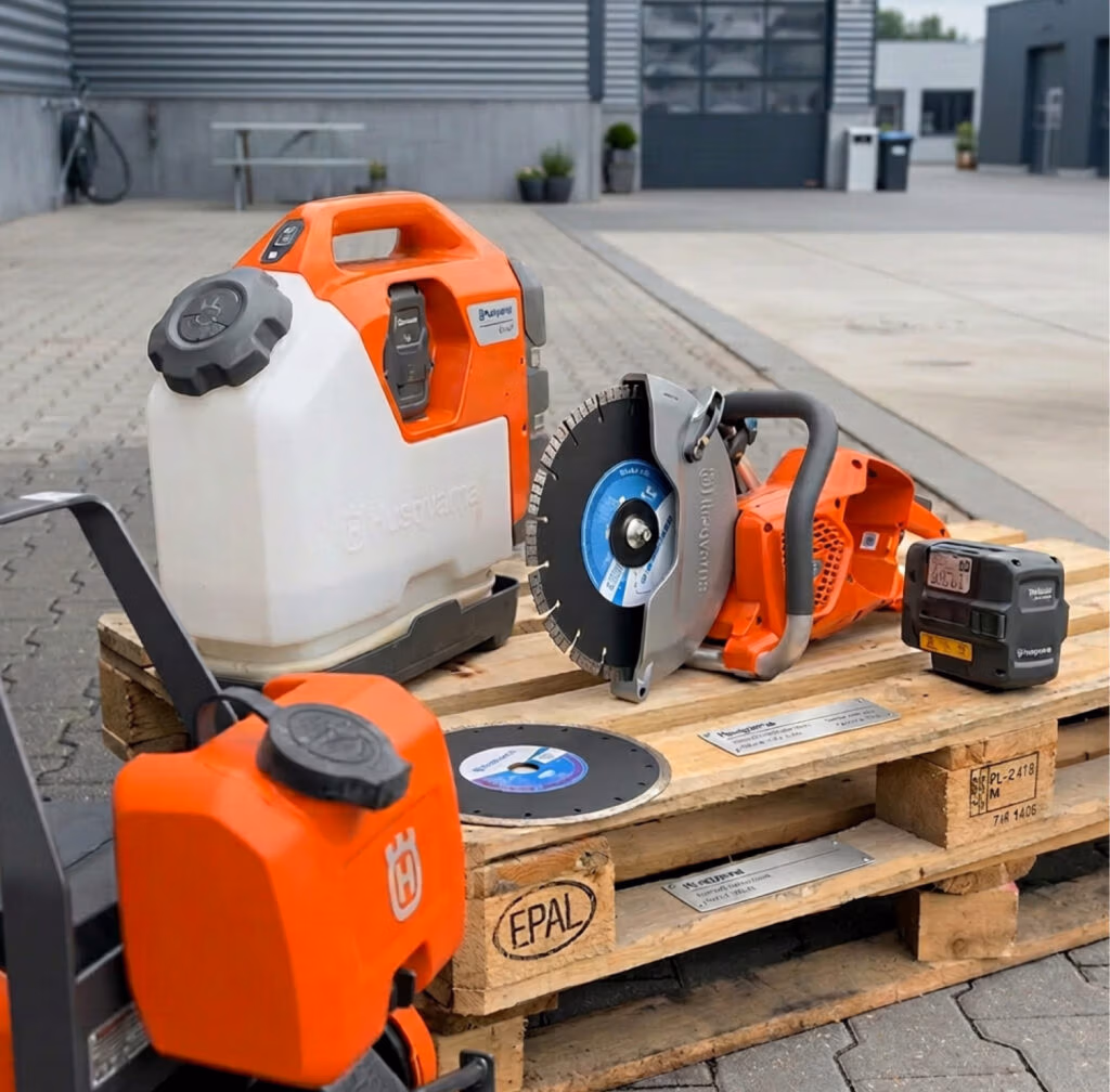 Op houten pallets buiten een modern industrieel gebouw, op een verharde ondergrond, staan een elektrische zaag, een waterreservoir, een accu en reservezaagbladen uitgestald. Deze selectie benadrukt de hoogwaardige gereedschappen van Thomaboor en Diamantboren, klaar voor professioneel gebruik.