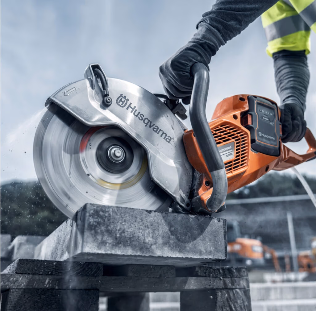 Iemand met handschoenen aan zaagt buiten met een oranje Husqvarna-motorzaag met hoogwaardige zaagbladen door een betonblok. Het draaiende zaagblad veroorzaakt stof, en op de wazige achtergrond zijn bouwvoertuigen te zien, wat de efficiëntie van professioneel gereedschap voor taken als beton zagen en diamantboren illustreert.