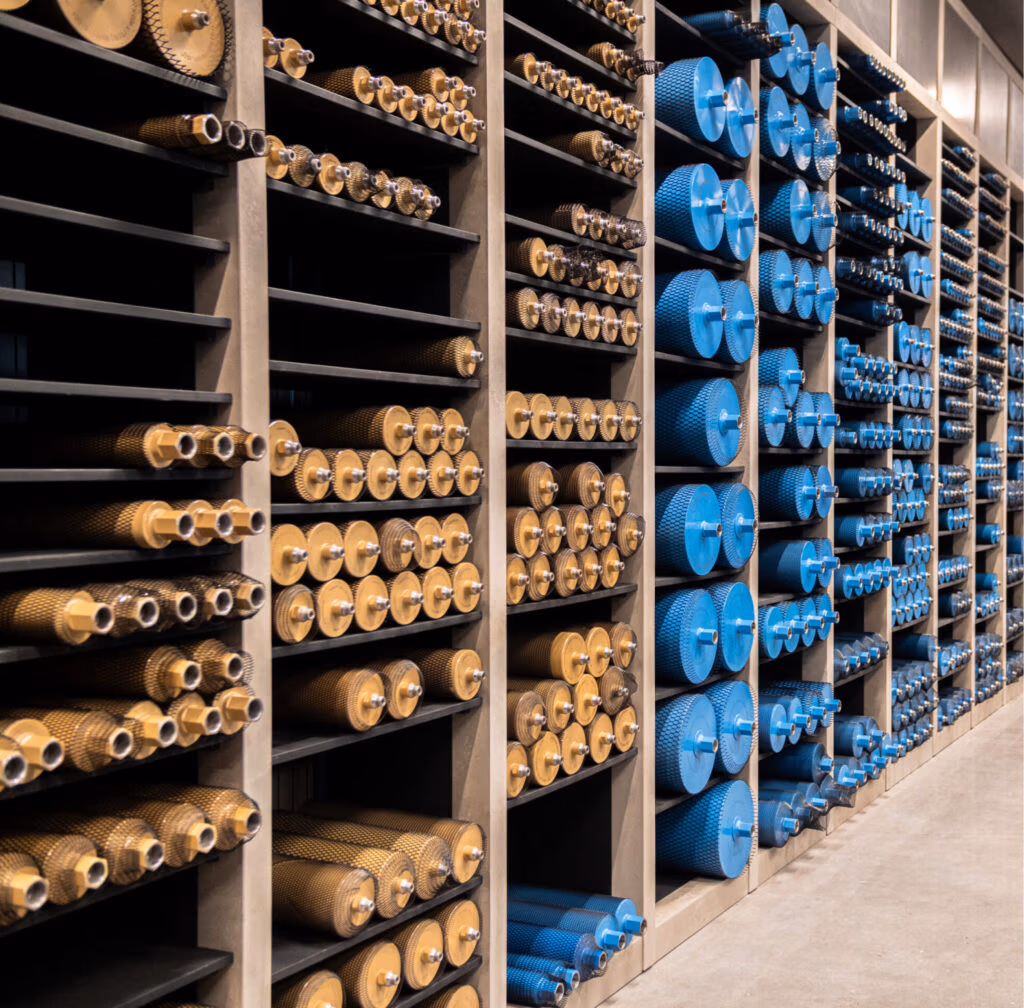 Rijen schappen vol met keurig geordende cilindrische metalen onderdelen; goudkleurige stukken aan de linkerkant en blauwe, gestructureerde onderdelen – zoals zaagbladen en diamantboren – aan de rechterkant, allemaal horizontaal opgeborgen in een werkplaats of industriële omgeving.
