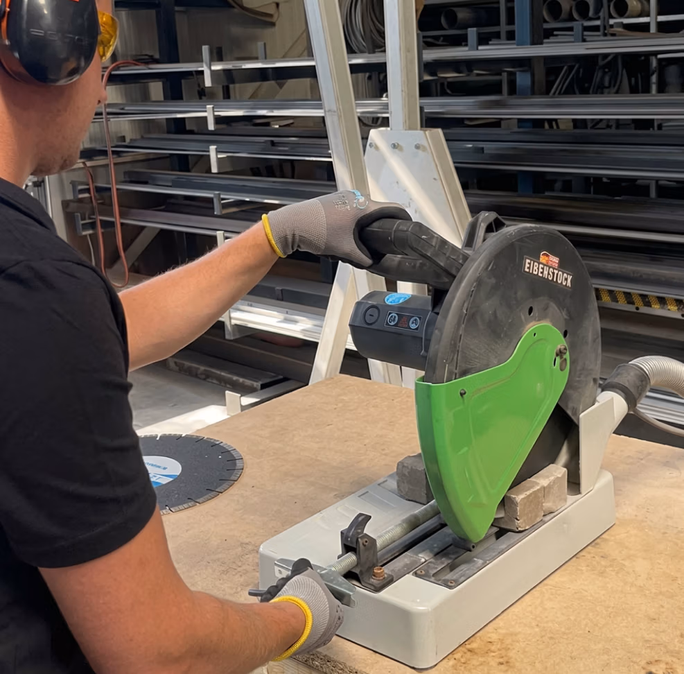 Een persoon die handschoenen en gehoorbescherming draagt, gebruikt een Eibenstock-motorzag, uitgerust met hoogwaardige zaagbladen, om een stuk metaal te zagen op een werkbank in een industriële werkplaats, waar ook gereedschap zoals diamantboren en Thomaboor veelvuldig wordt gebruikt.