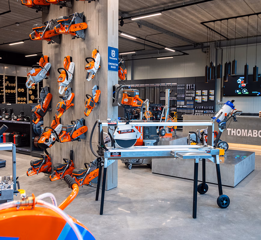 Een modern interieur van een Thomaboor-winkel met aan de muur meerdere oranje kettingzagen, terwijl op de tafels en planken op de achtergrond diverse zaagbladen en elektrisch gereedschap zijn uitgestald.