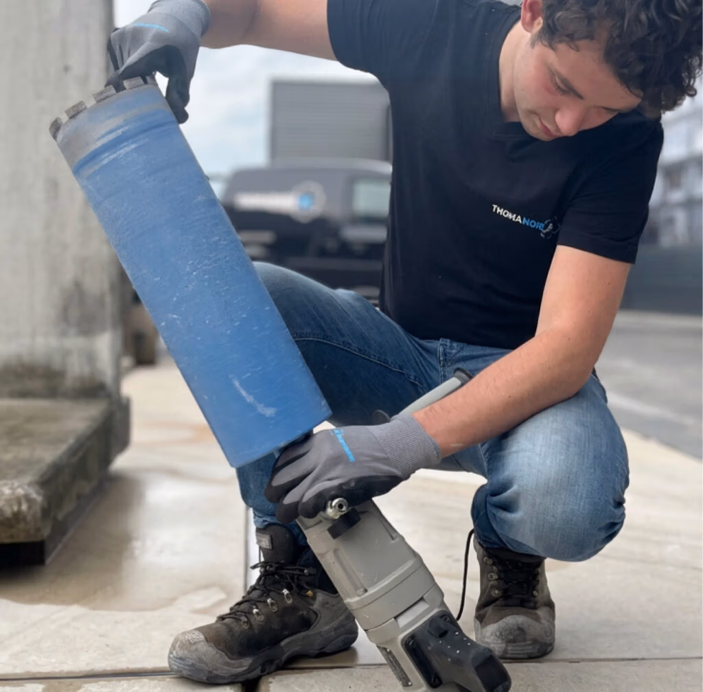 Een persoon in werkhandschoenen en een spijkerbroek knielt op een betonnen ondergrond en maakt zich klaar om buiten een groot blauw cilindervormig gereedschap te gebruiken, omringd door zaagbladen en diamantboren.