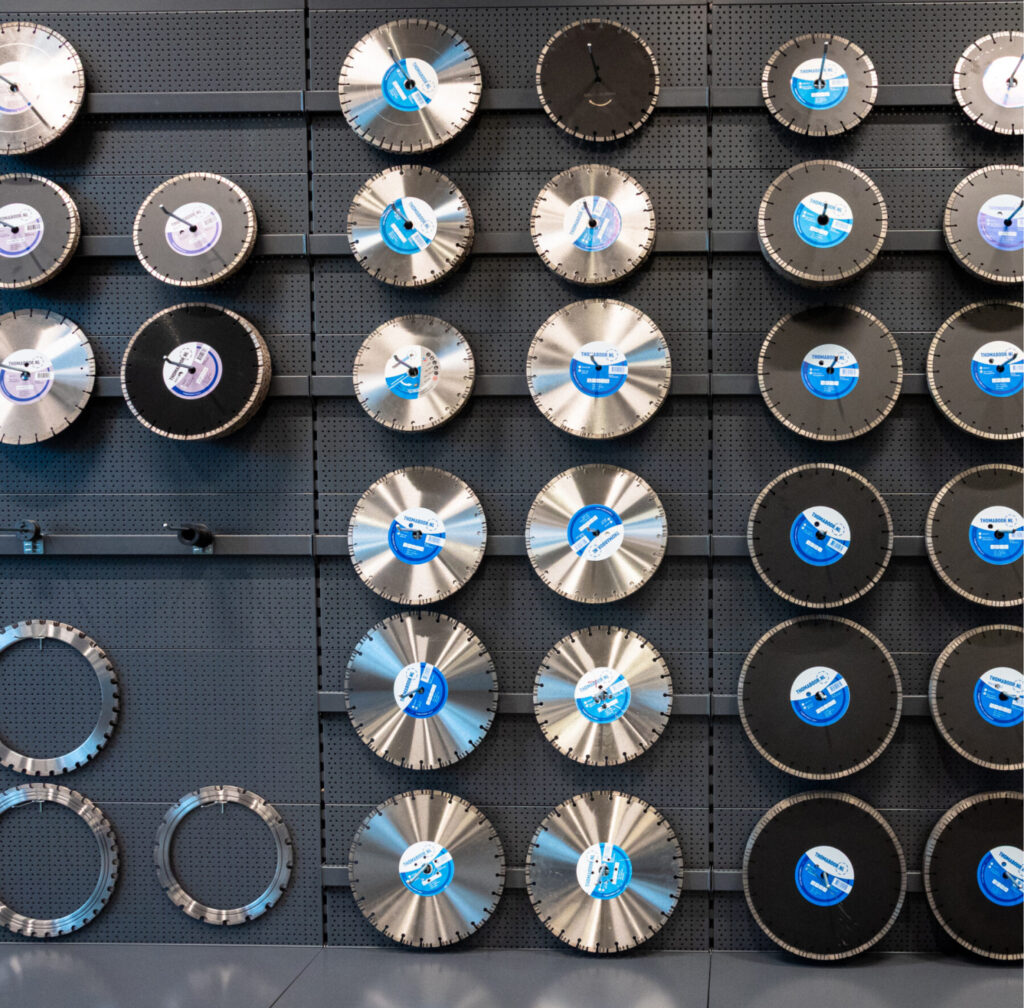 Cirkelzaagbladen en zaagbladen in diverse maten worden in een winkel of werkplaats tentoongesteld op een zwarte perforatieplaat, waarbij sommige zaagbladen zijn voorzien van blauwe etiketten en andere zijn afgedekt met zwarte beschermhoezen. Tussen het gereedschap kan ook een selectie diamantboren te vinden zijn, waardoor deze presentatie ideaal is voor professionals die op zoek zijn naar hoogwaardig zaaggereedschap.