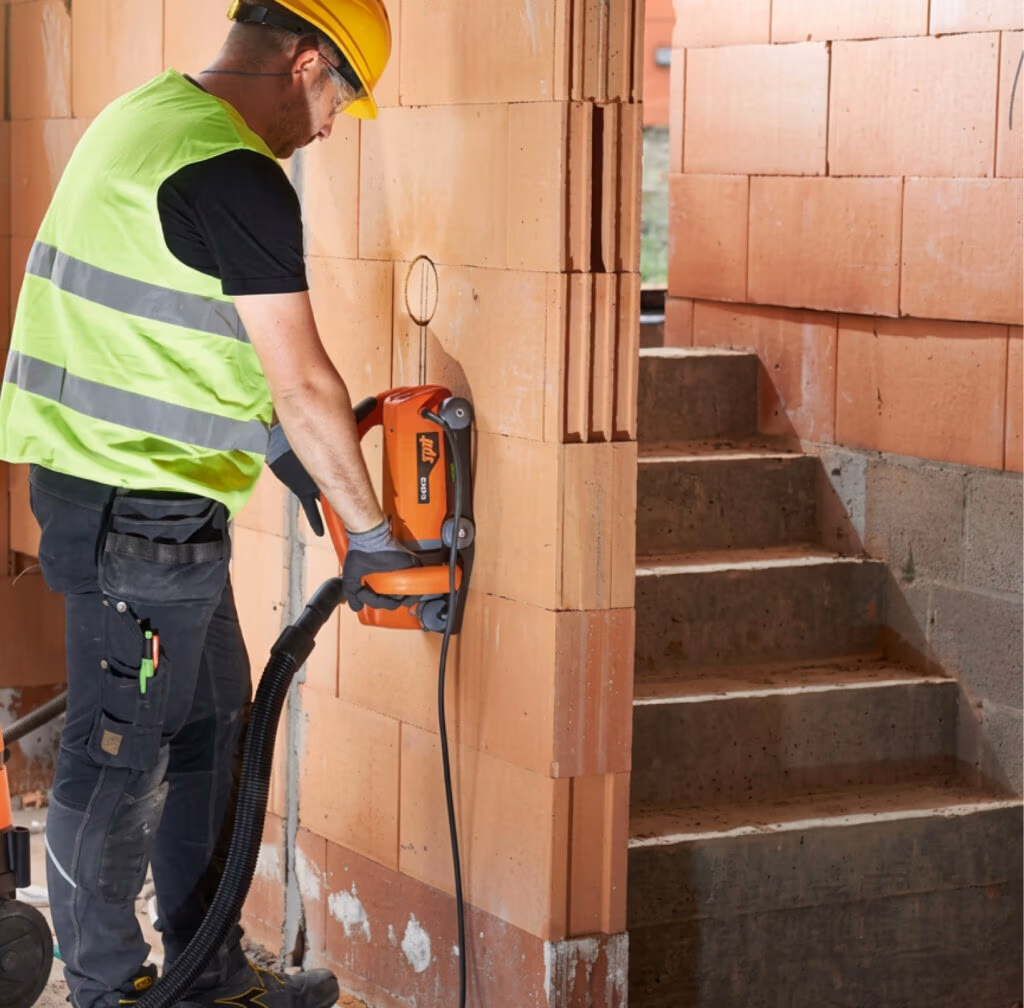 Een bouwvakker in een veiligheidsvest en met een helm op gebruikt een Thomaboor-elektrisch gereedschap met diamantboorbladen om sleuven te zagen in een bakstenen muur bij een trap in een gebouw in aanbouw.