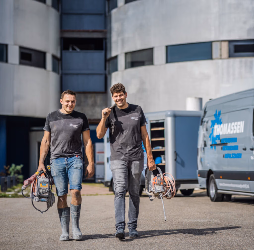Twee mannen in werkkleding lopen glimlachend voor een rond gebouw langs. Ze dragen gereedschap zoals een Thomaboor en diamantboren, en vlakbij staat een bestelbus geparkeerd. Het is een zonnige dag.