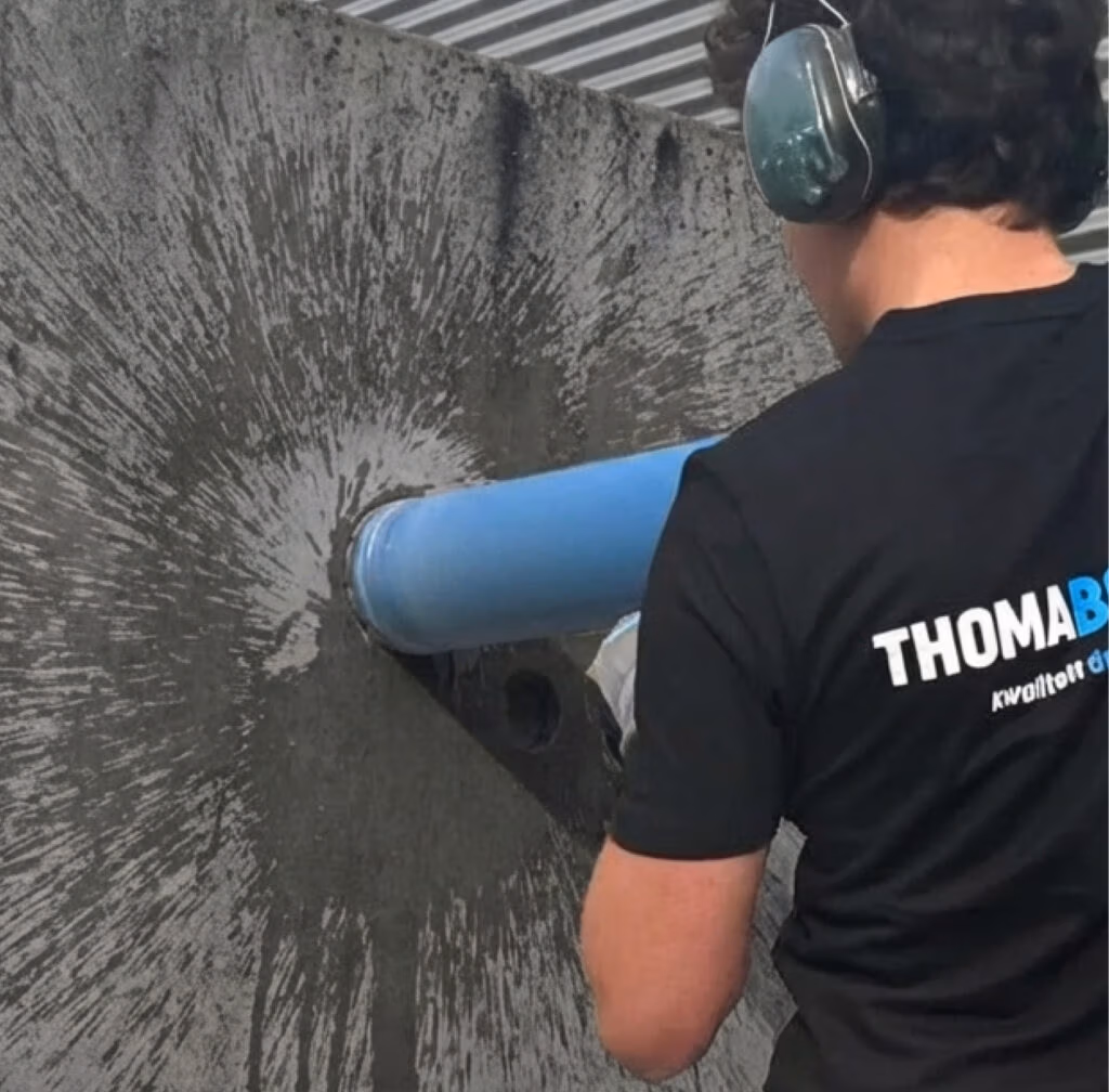 Met de juiste gehoorbescherming bedien een persoon een diamantboor om een rond gat in een betonnen muur te boren, waarbij stof naar buiten wordt geblazen. De persoon draagt een zwart shirt met op de rug het opschrift ‘THOMABOOR'. In deze industriële omgeving zijn onder het metalen plafond zaagbladen en andere apparatuur te zien.